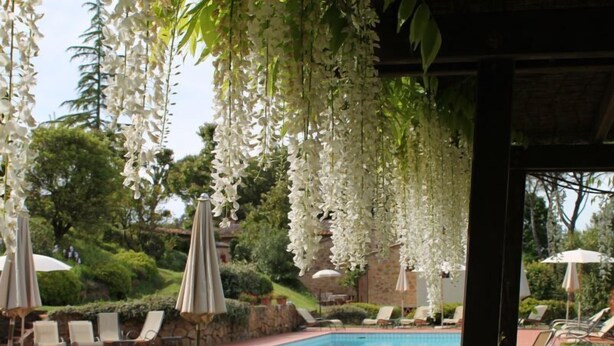 Zona de piscina con sillones reclinables y sombrillas, rodeada de flores blancas colgantes.