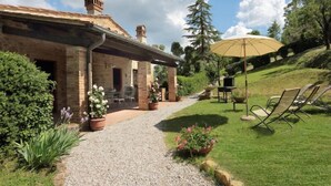 Un edificio de piedra con un patio cubierto, mobiliario exterior y un área verde con una sombrilla grande.