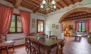 A dining area with a wooden table, chairs, and a fruit bowl. There are windows with curtains, a chandelier, and a fireplace. - POMARIUM - Main residence ground floor 135 m2 (Montaione)