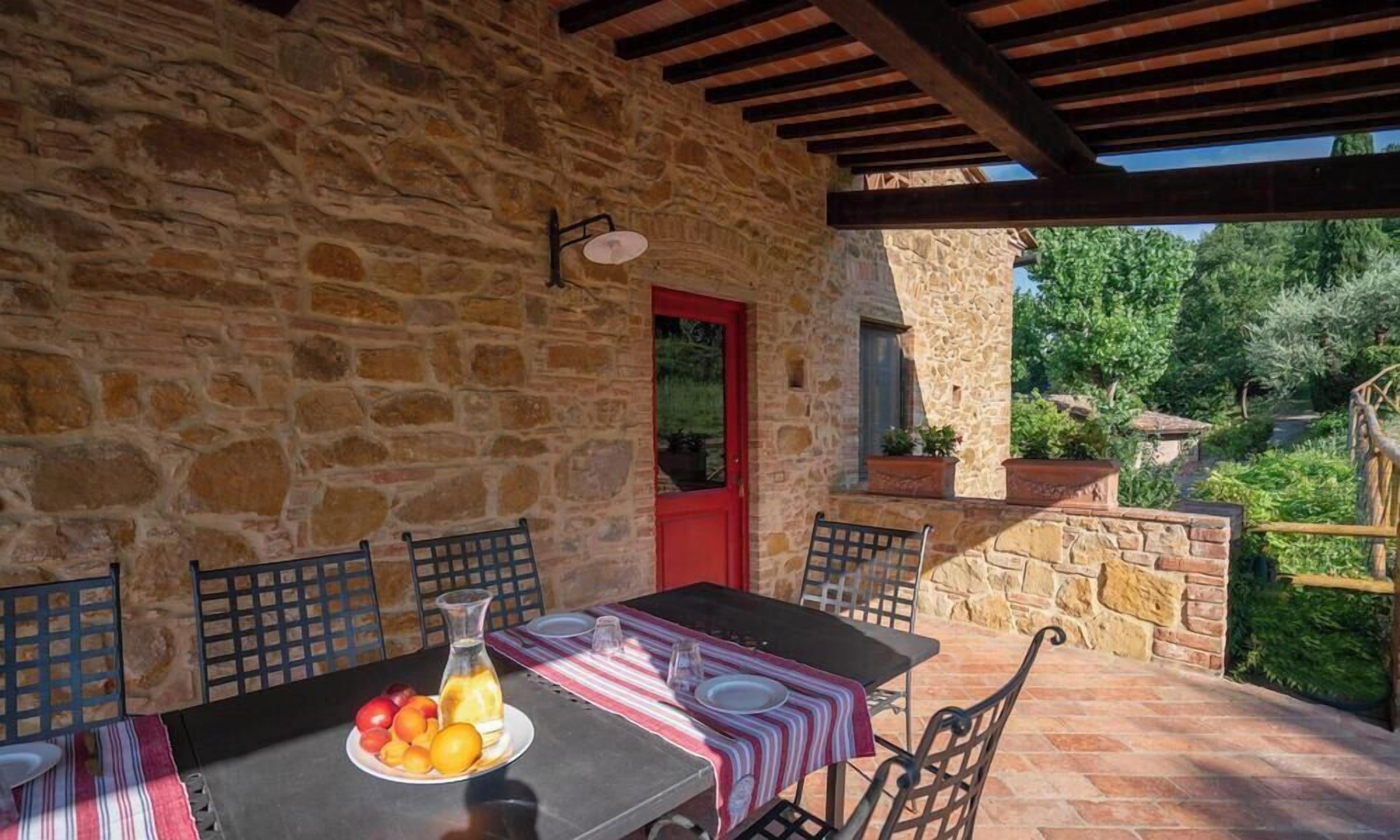 An outdoor dining area with stone walls, a red door, and a table set for two with a fruit bowl and a pitcher.