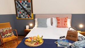 A hotel room with a bed, a chair, a basket of fruit, a book, and a handbag.