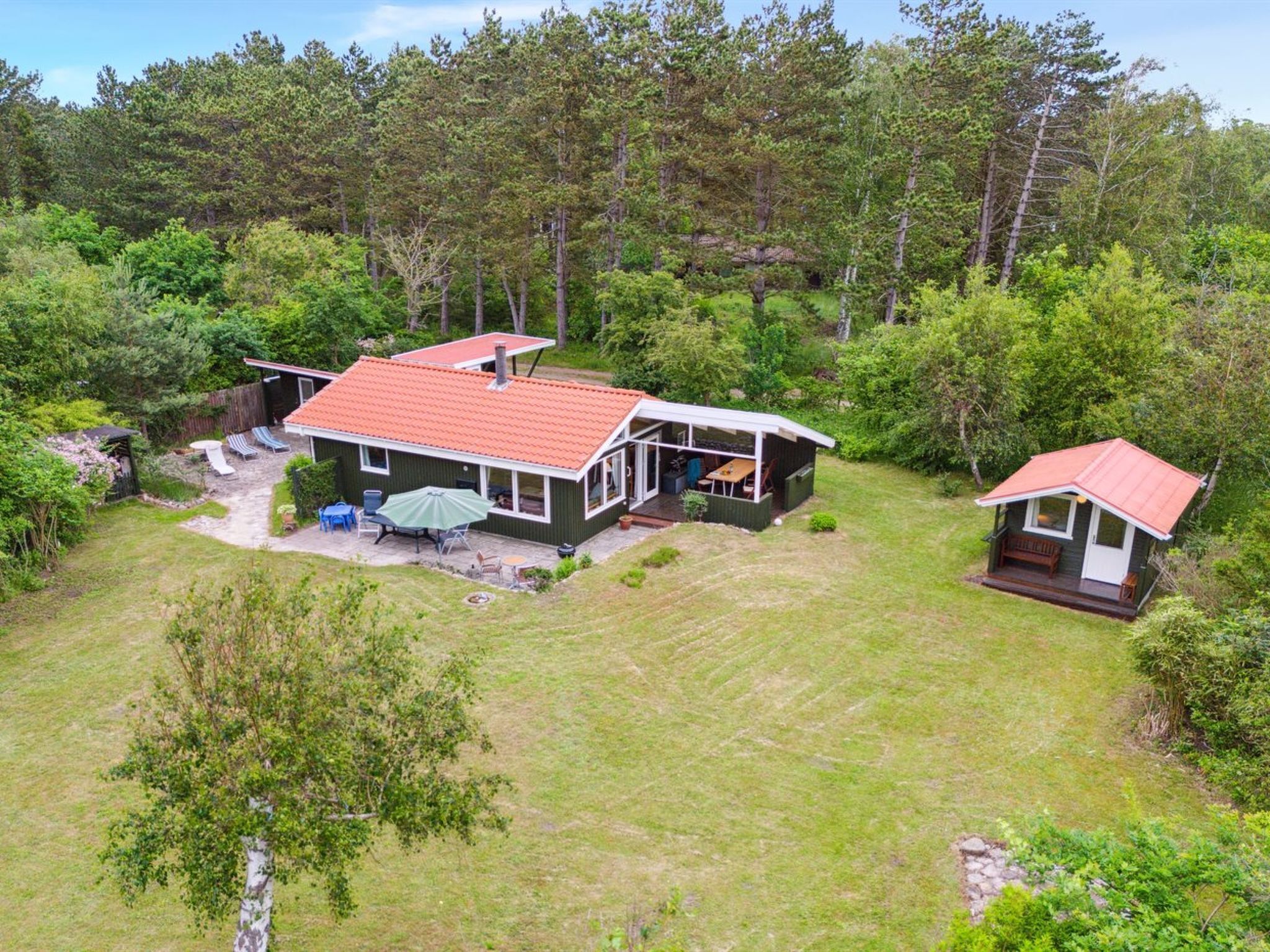 Ein zweistöckiges Haus mit rotem Dach, einer Terrasse mit Gartenmöbeln und einer kleinen Holzterrasse.