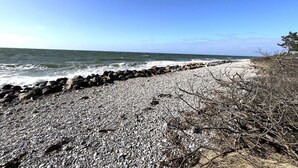 Uma praia rochosa com ondas quebrando na costa, céu claro e uma faixa de rochas separando a praia do oceano.