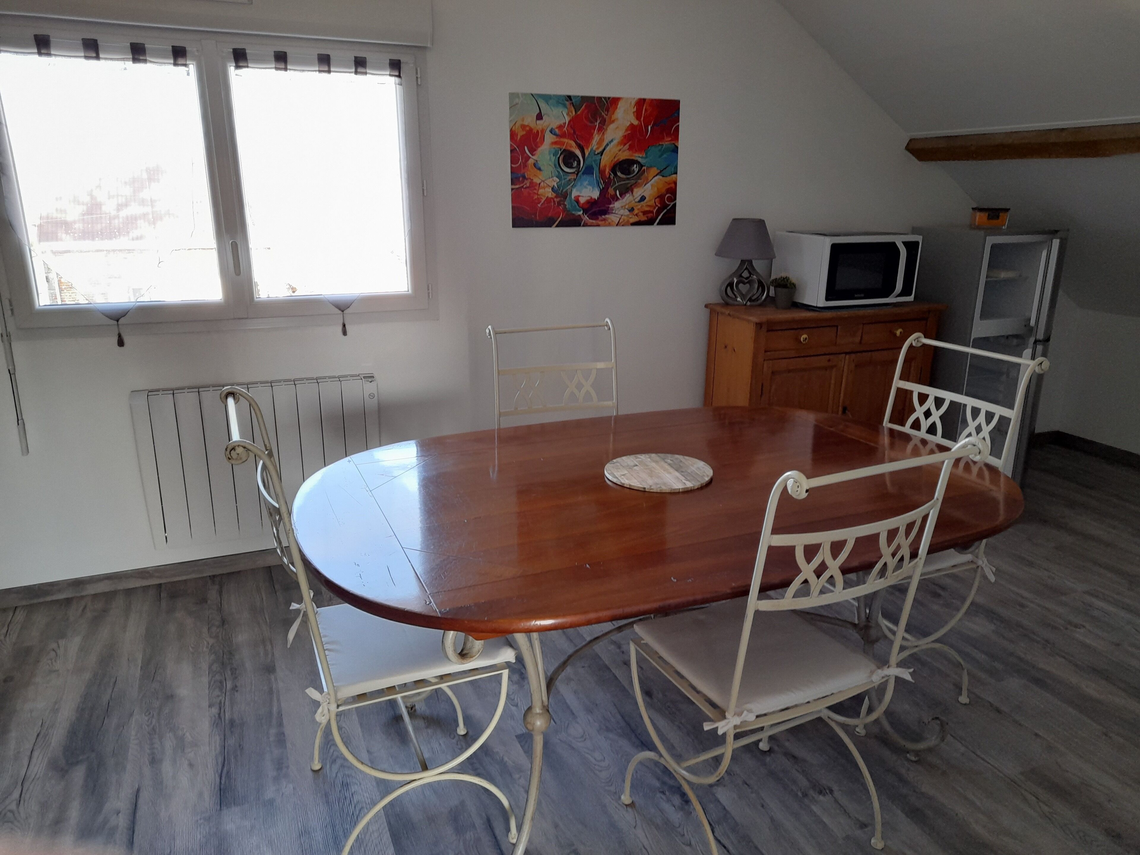 Comedor con mesa de madera y sillas de metal blanco, un radiador, un cuadro, un microondas y un armario.
