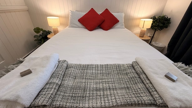 A neatly made bed with white linens, two red pillows, a grey and white woven throw, and two white towels with a small tag on each.