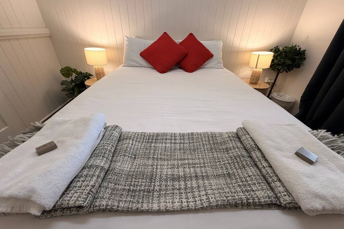 A neatly made bed with white linens, two red pillows, a grey and white woven throw, and two white towels with a small tag on each.