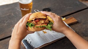 A person holding a burger with a drink in the background.