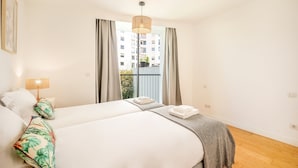 A hotel room with two beds, a balcony with curtains, a hanging light fixture, and a view of buildings. - GuestReady - One prime stay in Campolide (Lisbon)