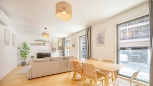 A modern living room with a sofa, dining table, chairs, and a large window. - GuestReady - One prime stay in Campolide (Lisbon)