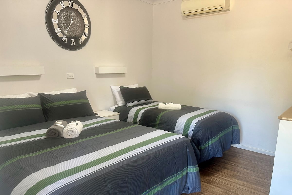 Habitación de hotel con dos camas, cada una con ropa de cama a rayas y almohadas, un reloj de pared y un equipo de aire acondicionado.