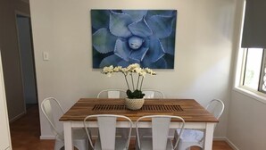 A dining area with a wooden table and white chairs, a vase with white flowers, and a large blue artwork on the wall. - Longreach Apartments (Longreach)