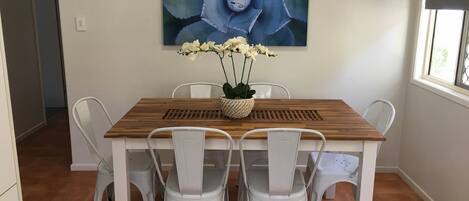 Un comedor con una mesa de madera y sillas blancas, un jarrón con flores blancas, y una obra de arte azul grande en la pared.