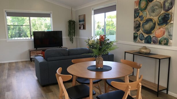 A living room with a grey sofa, a round wooden dining table with four chairs, a flat-screen TV, and a painting on the wall. - Longreach Apartments (Longreach)