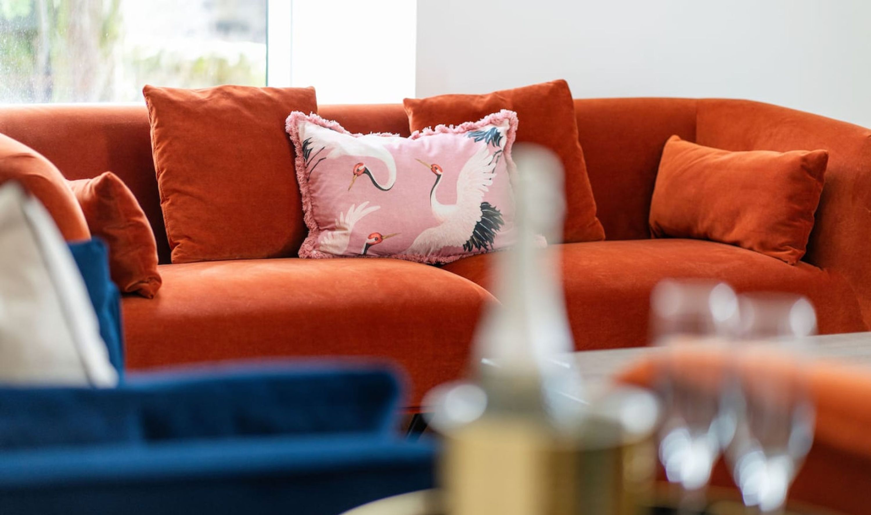 Een woonkamer met een oranje bank, een roze kussen met een vogelprint en een blauwe stoel.