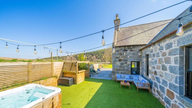 A stone building with a stone chimney, a wooden deck, a hot tub, and a seating area with cushions. - Kirkton Family Farmhouse With Hot Tub Alford Aberdeenshire (Alford)