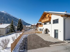 A modern building with a wooden balcony, a paved driveway, and a snow-covered landscape with mountains in the background.