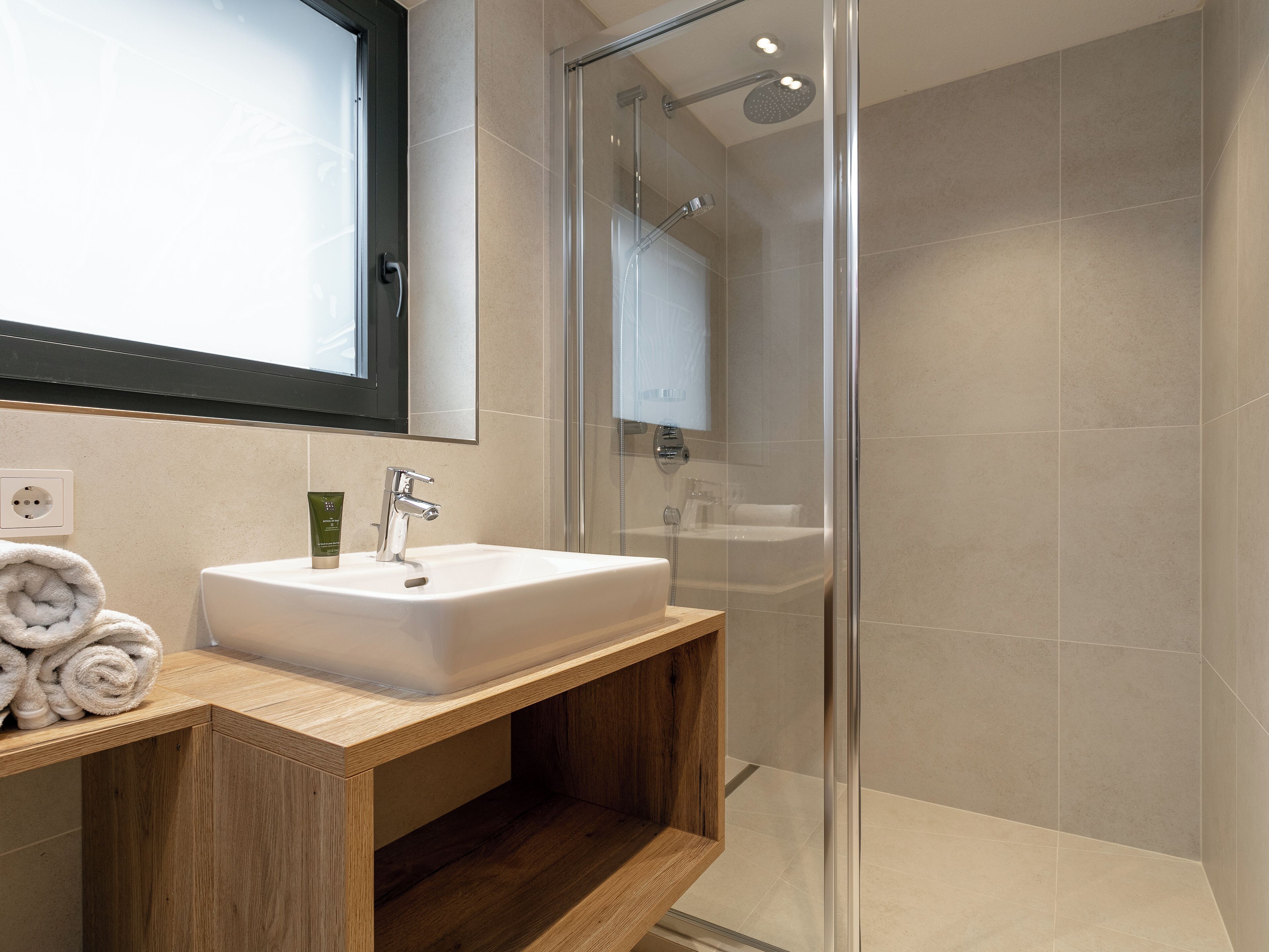Un baño moderno con lavamanos blanco, tocador de madera y zona de ducha con cerramiento de vidrio.