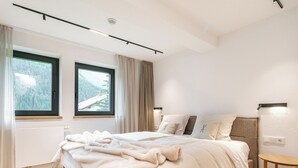 A modern bedroom with a large bed, two windows, a bedside table, and a view of a forested area.
