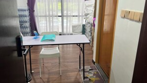 Une petite pièce avec une table, une chaise et une fenêtre ornée de rideaux violets. Des piles d’objets blancs sont disposées contre le mur, et une pancarte est accrochée à la porte.