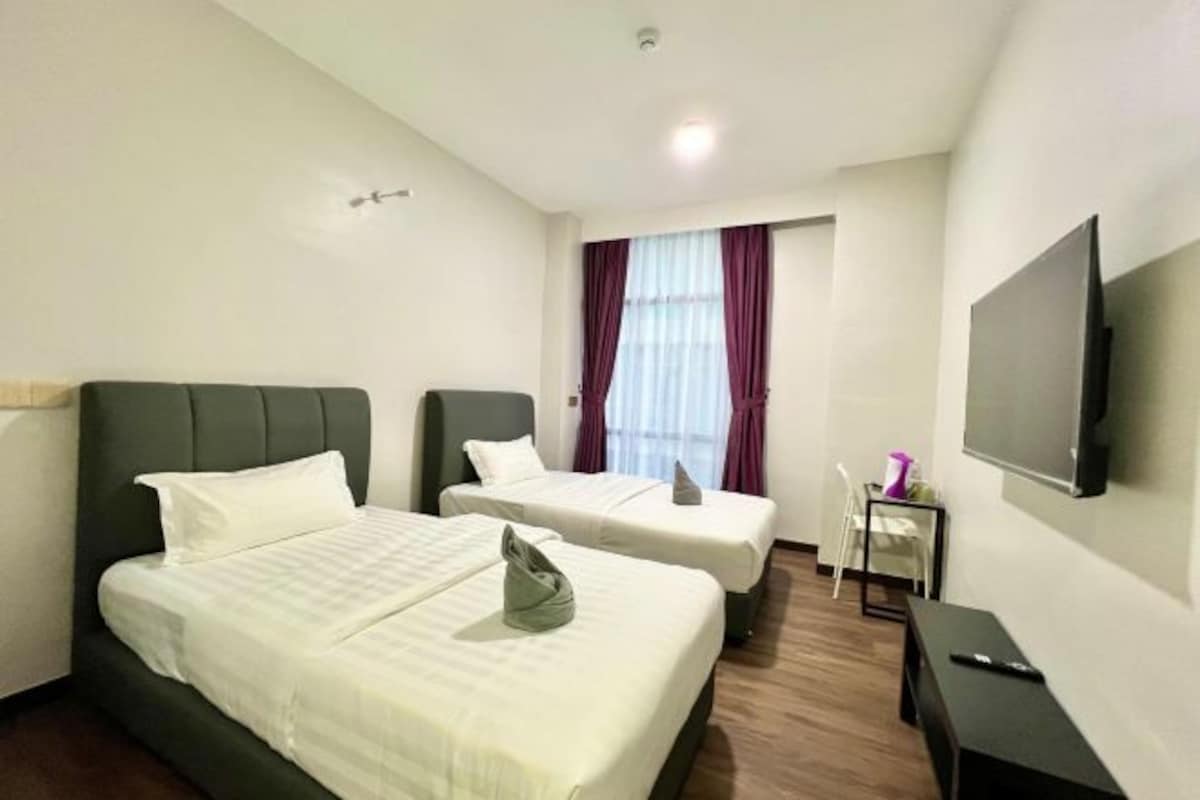 Habitación de hotel con dos camas, un televisor, una mesita pequeña y una ventana con cortinas.
