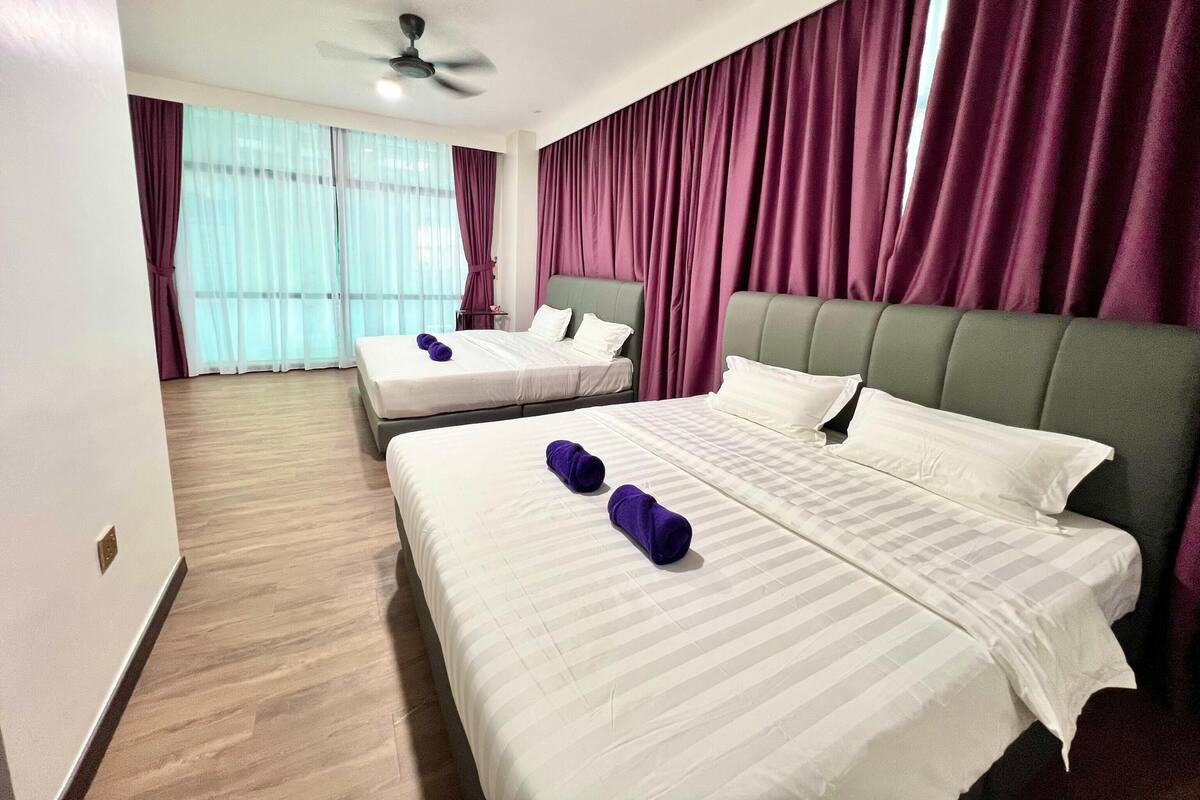 Une chambre d’hôtel avec deux lits, un ventilateur de plafond et de grandes fenêtres ornées de rideaux violets.