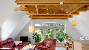 Un vaste espace de vie doté d’un haut plafond, d’un téléviseur, d’un canapé en cuir rouge, d’une table à manger avec des chaises et de grandes fenêtres offrant une vue sur la verdure.
