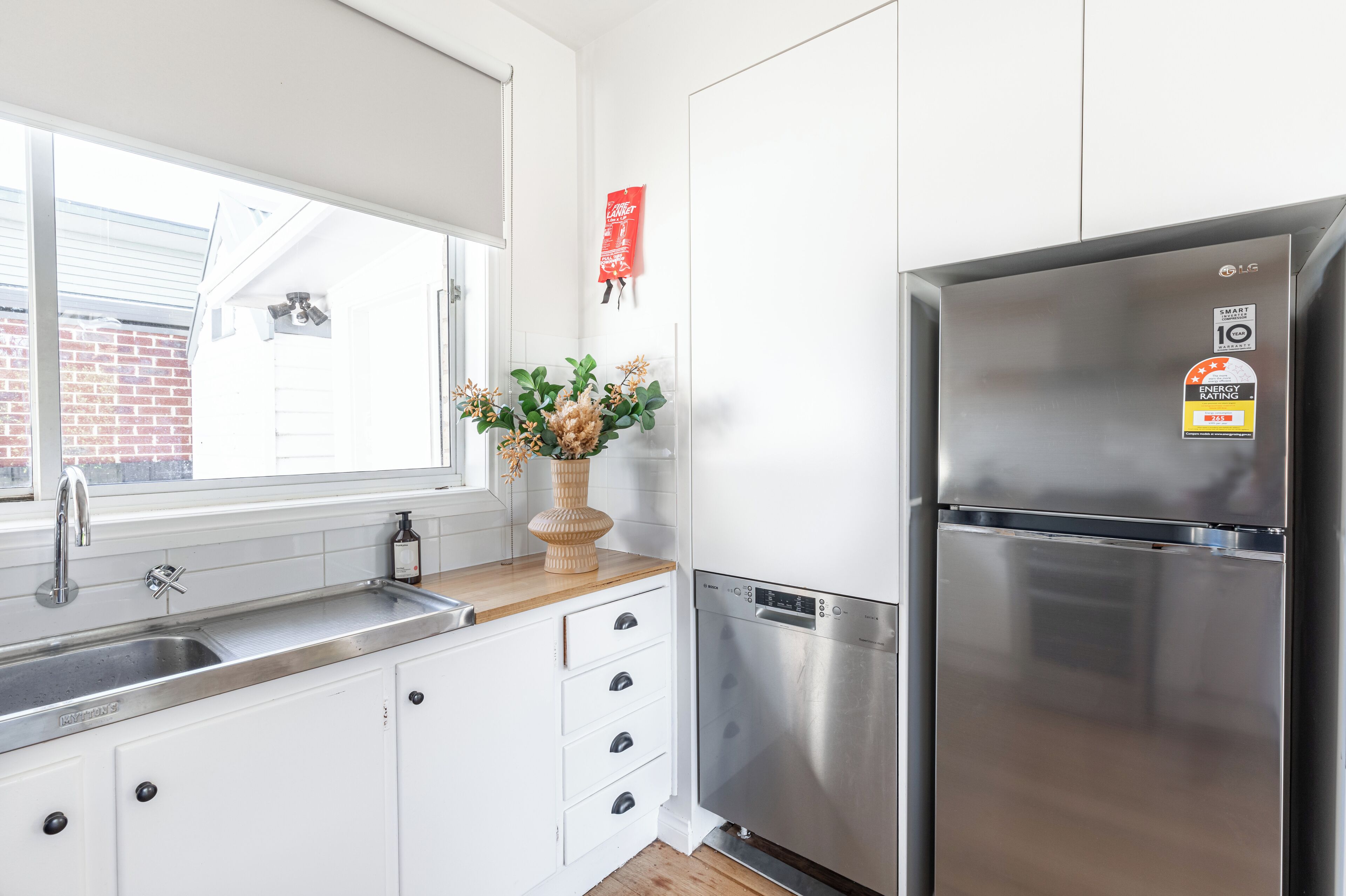 A modern kitchen with white cabinets, a stainless steel refrigerator, a built-in dishwasher, and a window with a view of a brick building.