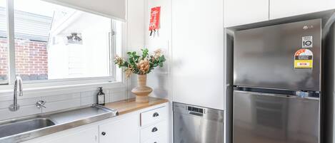 A modern kitchen with white cabinets, a stainless steel refrigerator, a built-in dishwasher, and a window with a view of a brick building.