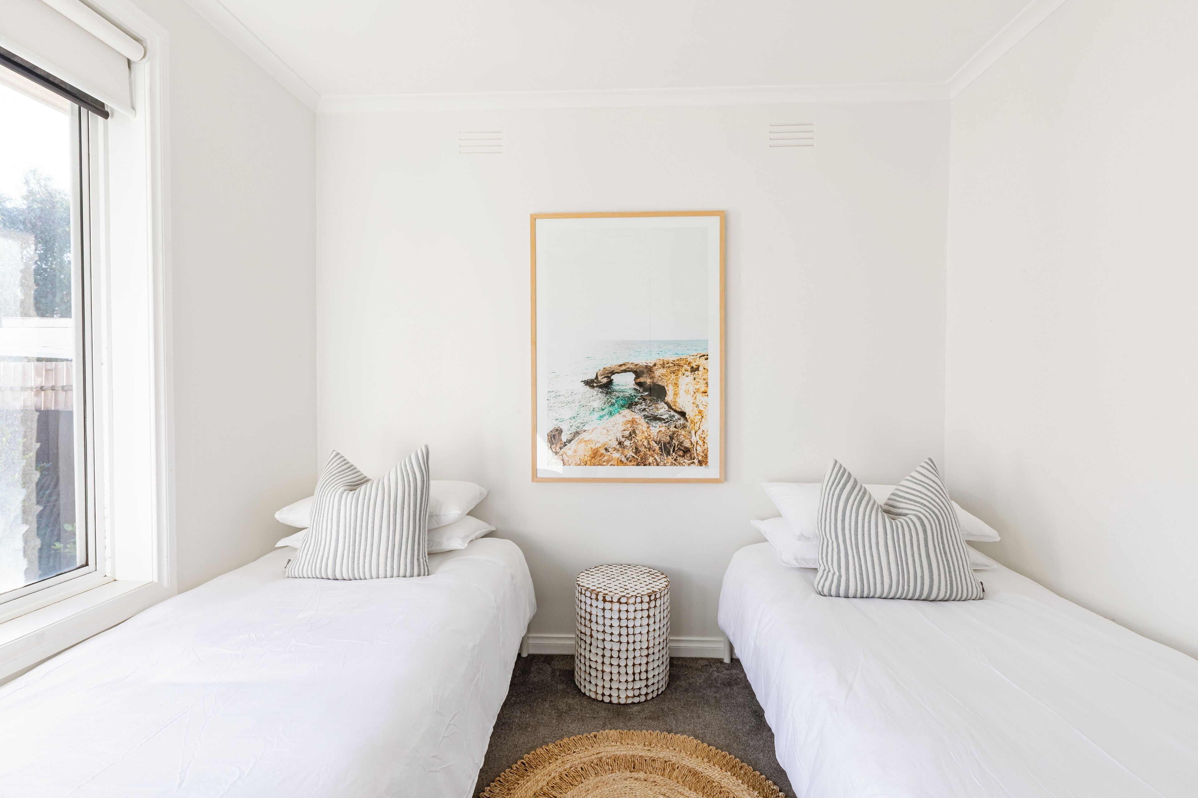 Une chambre avec deux lits, un tableau encadré au mur, un petit pouf rond et un tapis tissé.