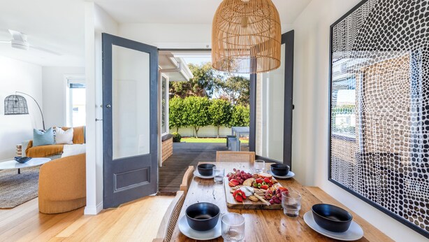 Un coin repas avec une table en bois dressée pour quatre personnes, un luminaire suspendu en osier et une vue sur un jardin, visible à travers une grande fenêtre.