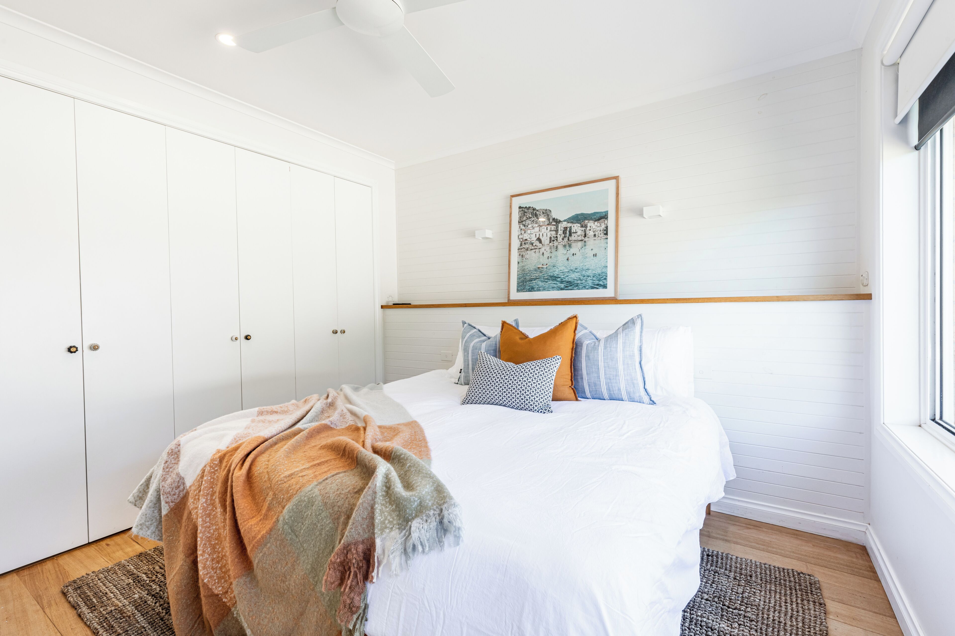 A bedroom with a large bed, white walls, a ceiling fan, a framed picture, and a window.