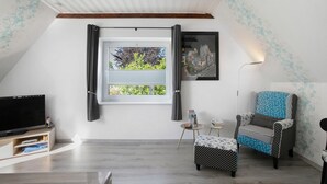 A room with a TV, a window with curtains, a chair, a small table, and a lamp. - Apartment 'De Lütte Moehl' with Natural Swimming Pond and Shared Terrace (Hollingstedt)