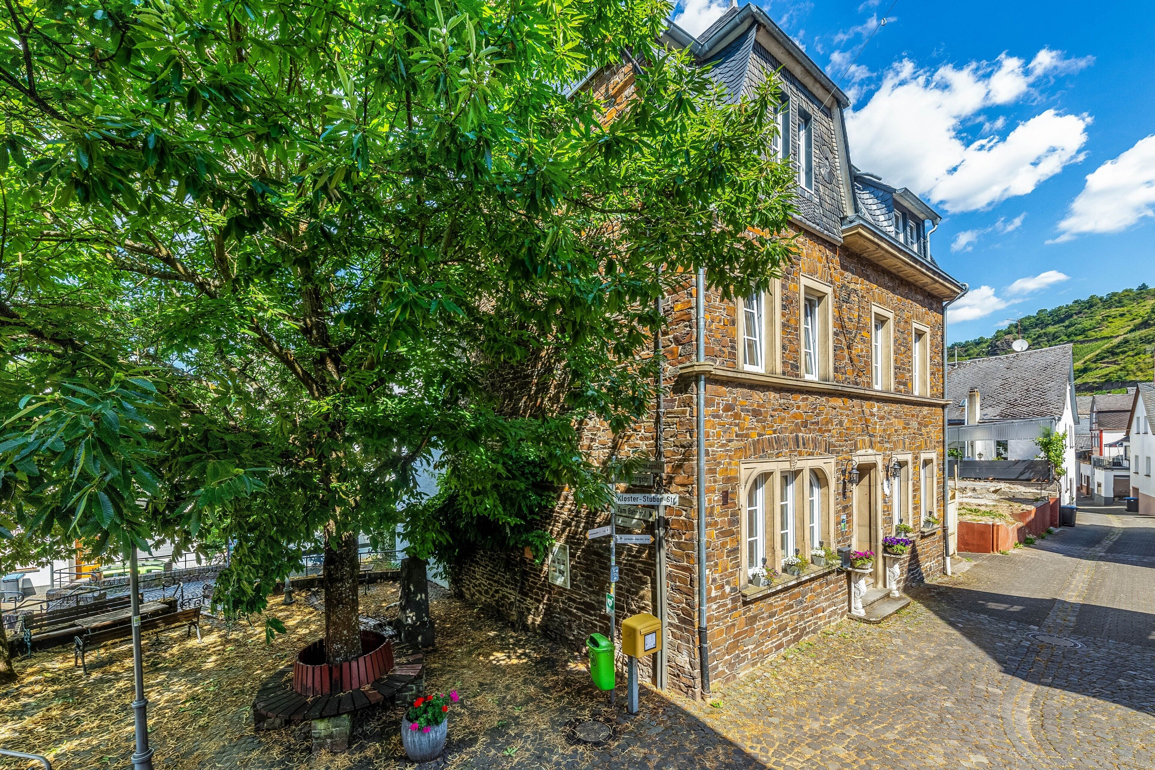 A stone building with a sloped roof, a large tree in front, and a cobblestone street.
