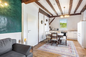 A cozy room with a wooden ceiling, a dining table with chairs, a sofa, and a view of greenery outside.