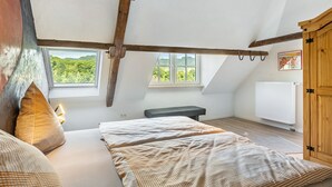 A bedroom with a sloped ceiling, wooden beams, a bed with patterned bedding, a bench, and a view of greenery through windows. - Spacious Holiday Apartment with Fireplace in Winzerhaus Schenk, with Mountain View and Wi-Fi (Neef)