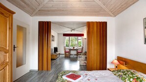 Une chambre d’hôtel avec un plafond en bois, un lit recouvert d’une literie à motifs floraux, une porte en bois et une vue sur un espace repas comprenant une table et des chaises.