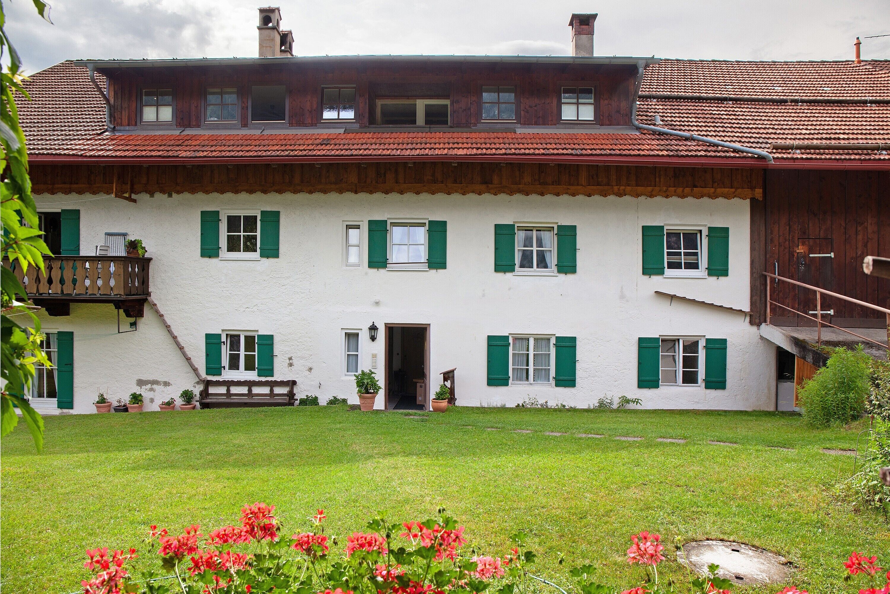 Ein zweistöckiges weißes Gebäude mit grünen Fensterläden, einem Balkon und einem kleinen Garten mit Blumen.