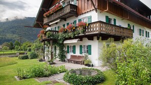 A traditional chalet with a balcony, a garden with a bench, and a mountain backdrop.