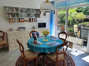 A dining area with a table set for two, a bookshelf, a television, and a view of the garden.