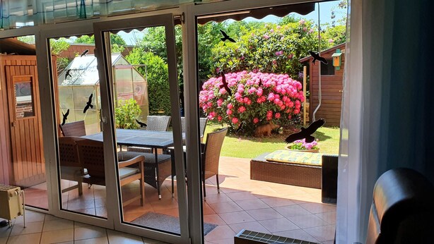 Une porte-fenêtre coulissante donne sur une terrasse extérieure aménagée avec une table, des chaises et un jardin fleuri.