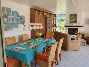 A dining area with a table set for four, surrounded by wicker chairs. A bookshelf and a television are visible in the background.