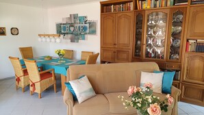 Un salon avec un canapé beige, une table à manger avec des chaises, une bibliothèque et une œuvre d’art murale.