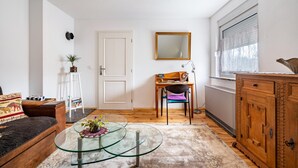 Ein Wohnzimmer mit Glastisch, einer Couch, einem Holzschrank, einem Sessel, einem kleinen Tisch mit Pflanze und einem Fenster mit Jalousien.