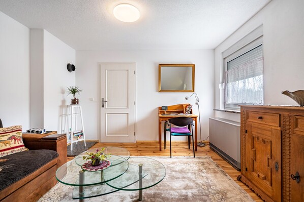 A living room with a glass coffee table, a sofa, a wooden cabinet, a chair, a small table with a plant, and a window with blinds.