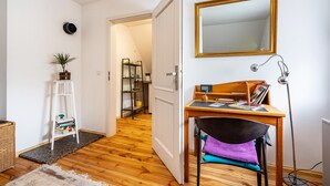 Ein Zimmer mit Holzboden, einem Schreibtisch mit Stuhl, einem Spiegel, einer Lampe und einer Tür zu einem anderen Raum.