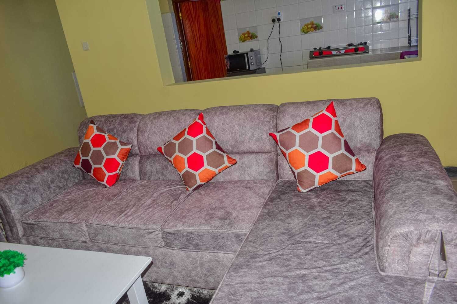 Un sofá gris con tres cojines estampados, una mesita blanca con una planta verde, y se ve una cocina a través de una ventana.