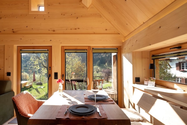 Un intérieur en bois avec une table à manger dressée pour deux personnes, de grandes fenêtres et une vue sur l’extérieur.