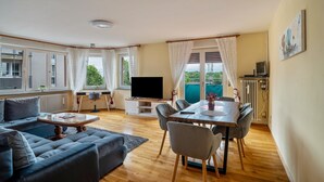 Un salon comprenant un canapé, une table basse, un téléviseur, une table à manger avec des chaises, et donnant sur un espace verdoyant.