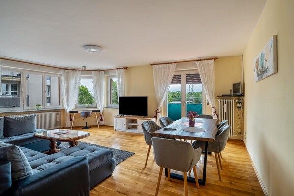 A living room with a sofa, coffee table, TV, dining table with chairs, and a view of greenery outside.