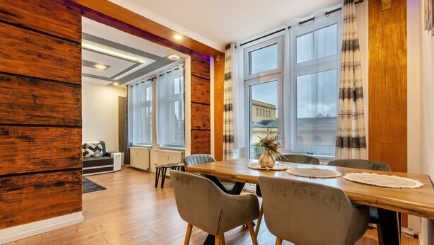 Un comedor con una mesa de madera, sillas grises y ventanas grandes con cortinas.